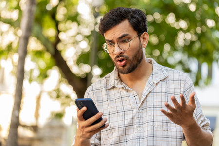 Young man using smartphone typing browsing, loses becoming surprised sudden lottery results bad news, fortune loss, fail. Guy standing on urban city street outdoors. Town lifestyles. Horizontalの写真素材