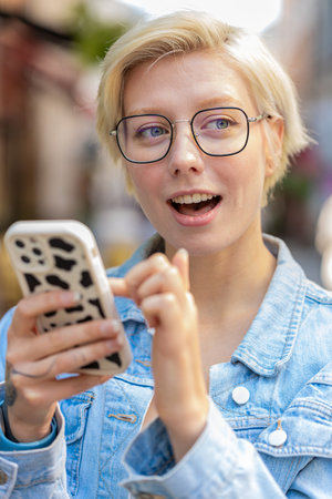 Caucasian young woman using smartphone typing texting social media messages e-mail looking for a way on map in mobile navigator app outdoors. Smiling traveler in glasses standing on city street.の写真素材