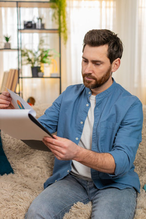 Man at home sits on the sofa, anxiously flipping through bills and documents searching for something crucial. Caucasian guy looks worried trying to find an important paper among financial statements.の写真素材