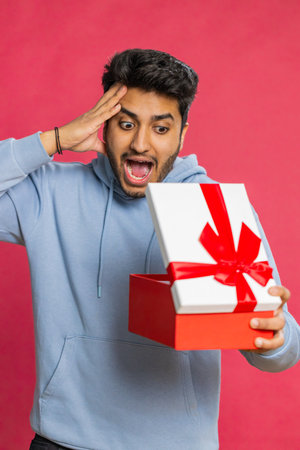 Excited man receiving a festive gift with a red bow opening the box with joy celebrating a surprise. Happy guy enjoying dream present smiling with excitement isolated on red backgroundの写真素材