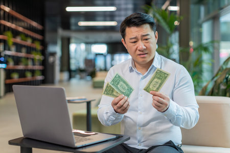 Depressed Asian businessman feeling worried about financial problem. Stressed man in shirt looking frustrated thinking of money debt, bad day, budget loss, bankruptcy sitting in modern office lobby.の写真素材