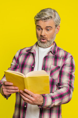 Mature man focuses intently on open book, revealing stunned curiosity perplexed gaze. Middle-aged guy on yellow background exhibits amazed wow reaction, hinting at new revelations hidden in textの写真素材