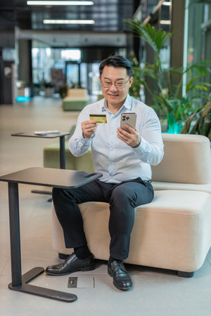 Happy businessman using bank credit card and smartphone for online shopping payments in modern office lobby. Business transaction. Smiling guy purchase with cellphone E-banking app service.の写真素材