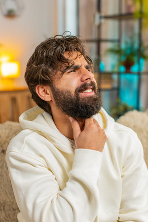 Upset young man in hoodie holds hand over sore throat. Painful neck and frowning, thyroid disorders, suffering sore throat, tonsils inflammation. Guy male sitting on sofa at home room.の写真素材