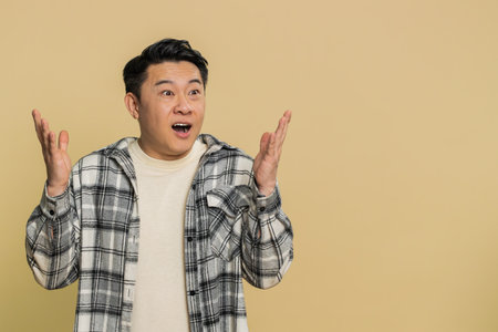 Excited amazed winner Asian man raising hands in surprise looking at camera with big eyes shocked by sudden victory celebrating success achievement good news. Guy on beige backgroundの写真素材