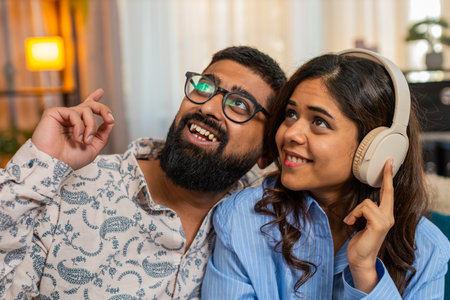 Indian couple man and woman sitting on home sofa, girl dances with headphones and boy joins her to...