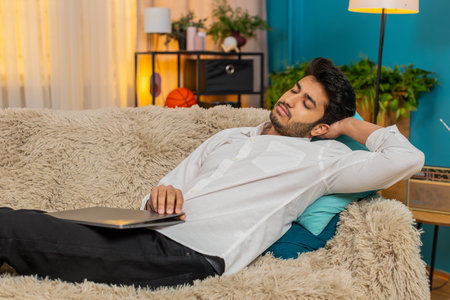 Young man at home, exhausted from work, falling asleep on the sofa with a laptop on his lap. Guy...