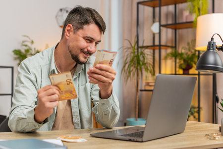 Planning budget. Rich happy man counting money euro cash using laptop calculate domestic bills at home table. Guy satisfied of income salary earnings, money for planned vacation gifts. Lifestyle.の写真素材