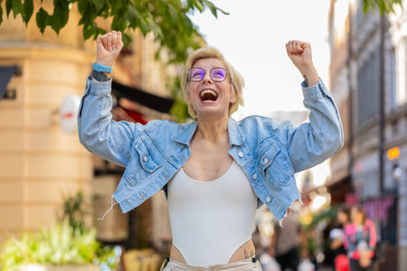 Excited surprised woman standing outdoors rejoices jumping with happiness feeling delight winning victory triumph win success result makes yes gesture. Lady traveler with blond hair on city street.の写真素材