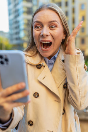 Happy shocked excited Caucasian mature businesswoman in coat mouth opened wow celebrate victory achievement, holding mobile phone good news win message rejoice success outside downtown city street.の写真素材