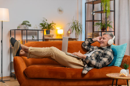 Happy relaxed Caucasian young man with gray hair in wireless headphones lying on home couch sofa choosing listening disco music while working on laptop netbook. Guy weekends activities entertainingの写真素材