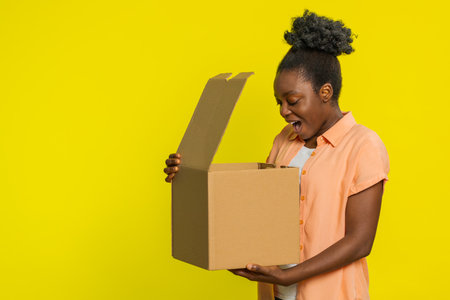 African American woman opens cardboard box, showing excitement joy while receiving a package. Black girl isolated on yellow background receive quick delivery and satisfaction with online purchaseの写真素材