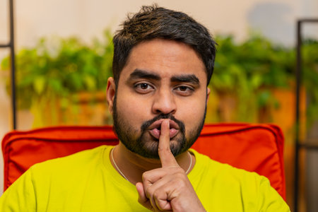 Shh be quiet please. Indian man presses index finger to lips makes silence hush gesture sign do not tells gossip secret. Bearded Arabian young guy at home modern apartment living room sitting on chairの写真素材