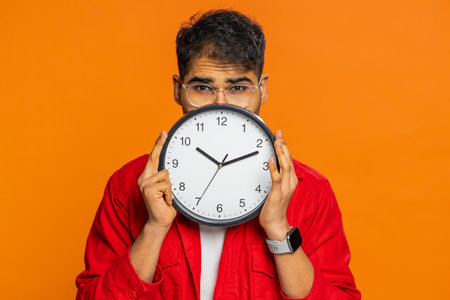 Serious strict Indian man holding office clock hiding behind, checking time on watch, running late to work, being in delay, deadline, obscuring face. Arabian guy isolated on orange background indoorsの写真素材