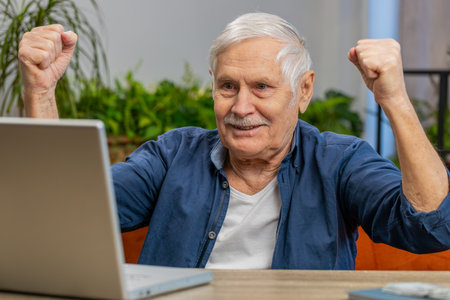 Happy senior old man at home couch with laptop scream in delight raise hands in triumph winner...