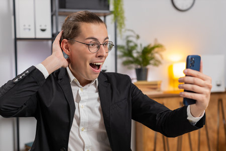 Oh my God, wow. Happy amazed young Caucasian businessman working on smartphone shocked by sudden victory, game winning, lottery goal achievement, good news. Stylish man freelancer guy at home office.の写真素材