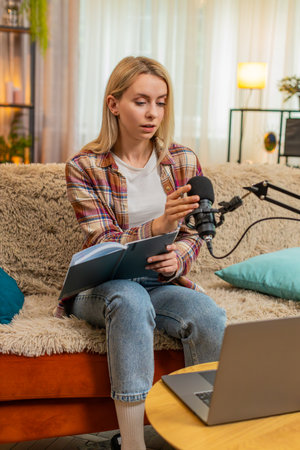 Blogger woman prepares to record podcast vlog on laptop while sitting on home sofa, adjusting mic and reading notes. Girl shows nervous energy and focus while testing sound and getting ready to speakの写真素材