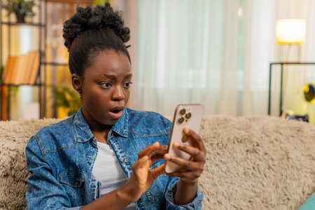 African American young woman suddenly opens her eyes wide in shock after reading unexpected and emotional social media news. Black girl stares at her phone, frozen by the intensity of what she sees.の写真素材