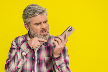 Mature man speaks on mobile phone with forceful tone, revealing anger agitation during tense talk quarrel. Middle-aged guy on yellow background shows argumentative stance, emphasizing heated dispute.の写真素材