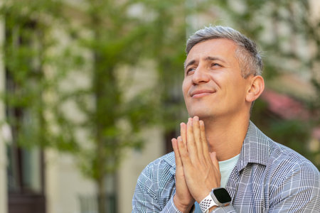 Grey-haired Caucasian mature man tourist praying with closed eyes to God asking for blessing help, forgiveness outdoor. Middle-aged guy clasping hands wishing luck in urban city street. Town lifestyleの写真素材