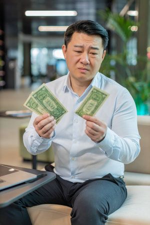 Depressed Asian businessman feeling worried about financial problem. Stressed man in shirt looking frustrated thinking of money debt, bad day, budget loss, bankruptcy sitting in modern office lobby.の写真素材