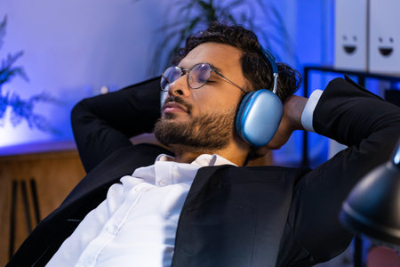 Happy smiling Indian businessman listening music on headphones taking break relaxing leaning on chair after working at office workplace. Male Hindu freelancer in formal suit puts hands behind headの写真素材