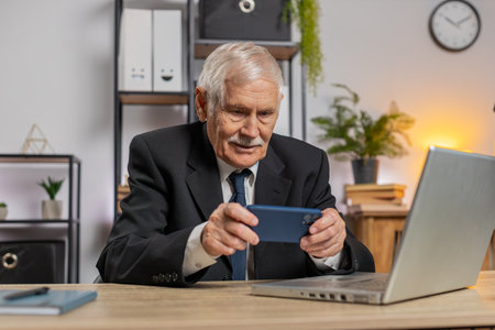 Caucasian senior businessman playing on smartphone exciting mobile application celebrate winning racing, shooter game at office desk workplace. Professional freelancer man enjoy good work of new app.の写真素材