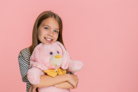 Young child girl gently hugs a plush toy bunny with love and happiness, smiling sweetly in joy, expressing warmth and emotional connection. Little teenager school kid isolated on pink background.の写真素材