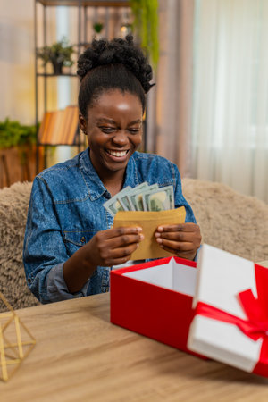 African American young woman sitting at table on home sofa happily checks envelope full of cash dollars received as a birthday gift. Black girl opens big gift box and smiles with joy and surprise.の写真素材