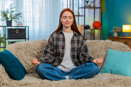 Young woman at home meditating in lotus pose on sofa with closed eyes, breathing deeply in full...