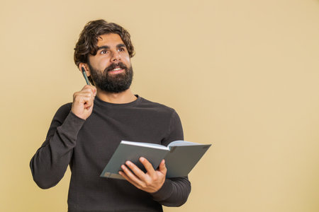 Thoughtful young Indian business man making notes, writing down thoughts with pen into notepad notebook diary, to do list, good idea. Arabian adult guy isolated on beige studio background indoorsの写真素材