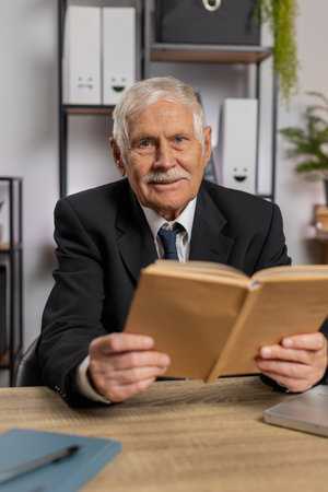 Bored Caucasian senior businessman reading book, literature, taking a rest after work. Grandfather freelancer guy in suit with laptop relaxing with head in hand at home office table. Business people.の写真素材