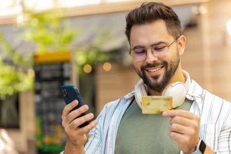 Caucasian young man using credit bank card and smartphone while transferring money, purchases online shopping, order food delivery, booking hotel room. Happy tourist guy on urban city street outdoors.の写真素材