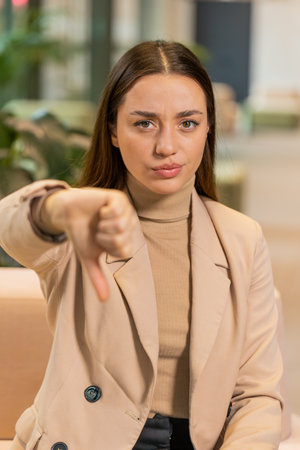 Dislike. Upset Caucasian businesswoman sitting in office lobby showing thumb down sign gesture, expressing discontent, disapproval dissatisfied bad work. Displeased serious freelancer in formals.の写真素材