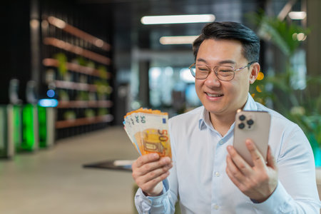 Successful happy rich mature businessman in shirt holding smartphone and counting euro bill money cash fan sitting in office lobby. Happy Chinese guy winning online lottery game. Calculating budget.の写真素材