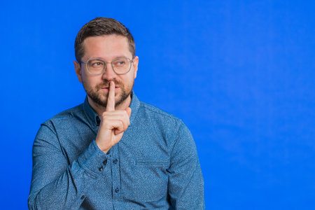 Shh be quiet please. Caucasian middle-aged man in glasses presses index finger to lips makes silence gesture sign do not tells secret, stop talk gossip, confidential privacy. Guy on blue backgroundの写真素材
