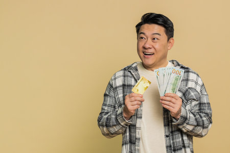 Happy Asian middle-aged man showing plastic credit bank card and money dollar cash advertising taking loan financial help for purchases online. Mature Chinese guy isolated on beige background indoorsの写真素材