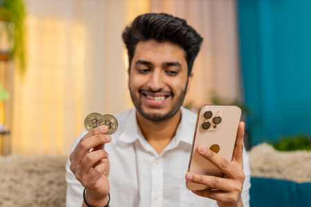 Indian man at home on sofa checking bitcoin wallet balance on smartphone, feeling excited slightly tense. Guy focused on cryptocurrency market fluctuations, reacting to financial changes in real timeの写真素材