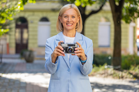 Smiling happy excited mature Caucasian businesswoman photographer in formal suit using vintage camera to take photos pictures of fascinating city. Travel, hobby concept. Lady employer on city street.の写真素材