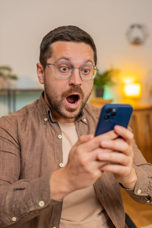 Oh my God, Wow. Caucasian young businessman using smartphone shocked by sudden victory game winning, lottery goal achievement, good news. Professional manager freelancer guy sitting in home office.の写真素材