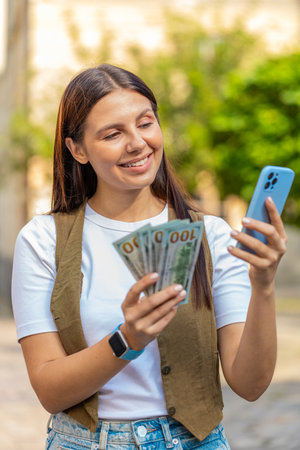 Happy young woman counting money dollar cash, using smartphone calculator app, satisfied of income for planned vacation gifts. Lovely girl standing in urban city sunny street. Town lifestyles outdoorsの写真素材