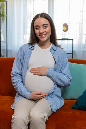Portrait of smiling future mother sitting on sofa touching pregnant belly in living room at home....