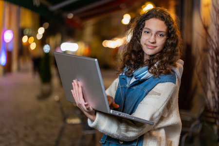 Portrait of smiling young girl working online distant job with laptop in city street, browsing website chatting outdoors. Caucasian woman tourist thinking typing in town at night. Lifestyles.の写真素材