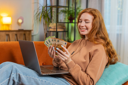 Planning budget. Rich happy young woman counting money euro cash using laptop calculate domestic bills sitting on sofa. Girl satisfied of income salary earnings saves money for planned vacation, giftsの写真素材