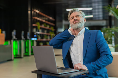 Tired Caucasian middle-aged businessman suffers from spine neck muscle pain working on laptop in office lobby. Overworking. Male entrepreneur man guy in suit attempt to relieve pain by massaging.の写真素材