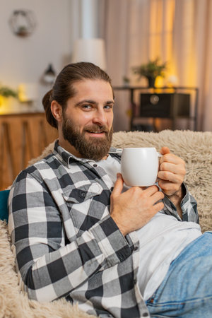 Young man resting lying at home, holding warm cup of coffee or tea with gentle grin and eased shoulders. Caucasian guy on sofa feels soothing quiet, appreciating short pause from busy tasks and noise.の写真素材