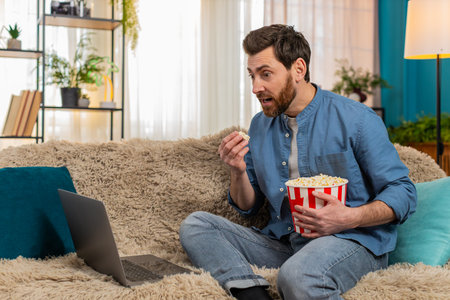 Young man at home enjoys popcorn while watching movie on laptop, fully immersed in scene. Caucasian guy laughing in excitement at an unexpected moment, reacting emotionally to the thrilling storylineの写真素材