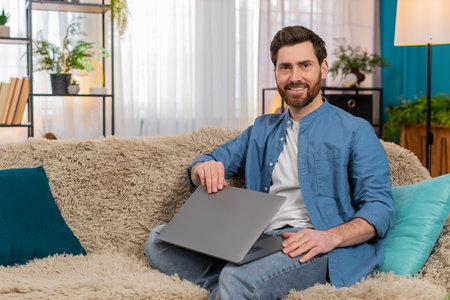 Young man at home smiling with satisfaction while closing laptop after completing work. Caucasian guy sitting on sofa, feeling accomplished, relieved, and happy about finishing tasks or projectsの写真素材