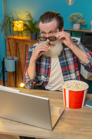 Caucasian elderly man freelancer taking break from work wearing 3D glasses eating popcorn and watching movie on laptop. Laughing grandfather sitting at home office desk having snacks during work breakの写真素材