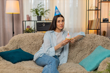 Lonely young woman at home sadly celebrating birthday alone holding cake with lit candles. Caucasian unhappy girl on sofa emotionally disappointed, no guests attending, feeling isolated and upsetの写真素材